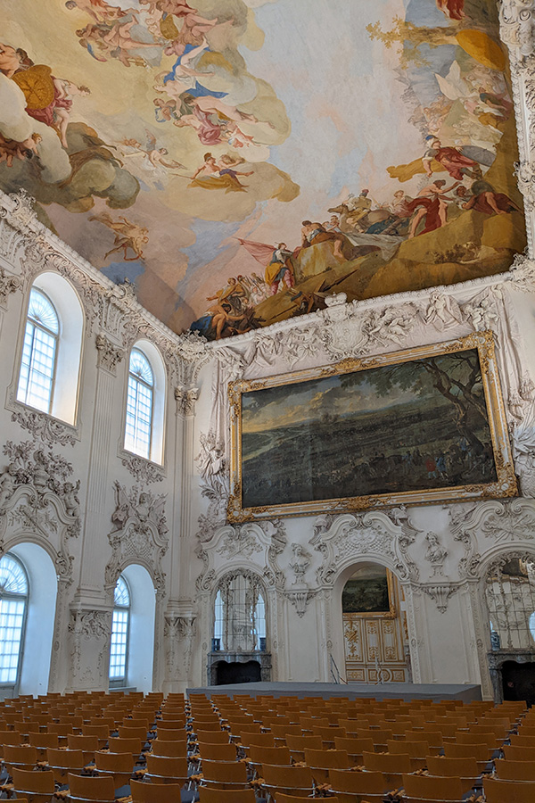 A large painting hangs below a detailed ceiling fresco in the Large Hall of Schleissheim Palace.