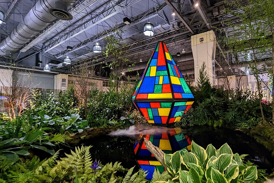 A glass pyramid stands on the edge of a misty pond in a lush landscape exhibit called In Search of Peace at the Philadelphia Flower Show 2023.