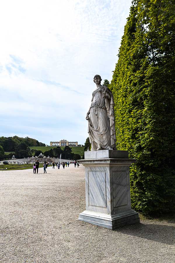 A stone statue stands outside Schloss Schönbrunn with the Gloriette in the distance upon the hill.