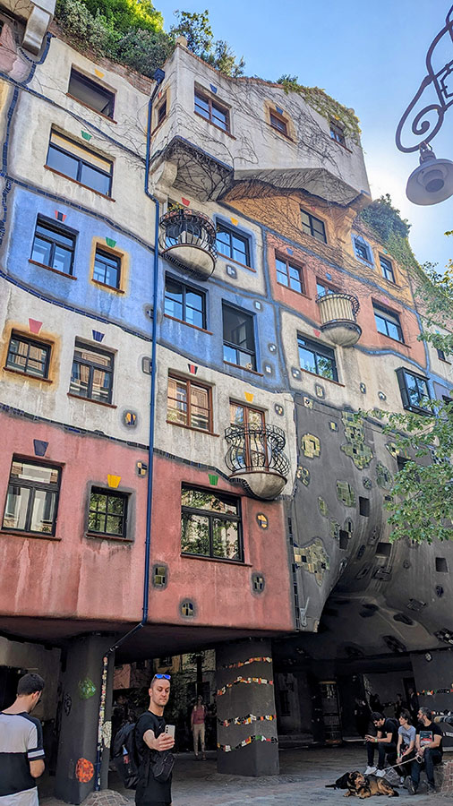 Portrait-oriented view of the colorful and eclectic exterior of the Hundertwasserhaus in Vienna, showcasing its irregularly shaped windows, uneven façade, and lush greenery on the rooftop.