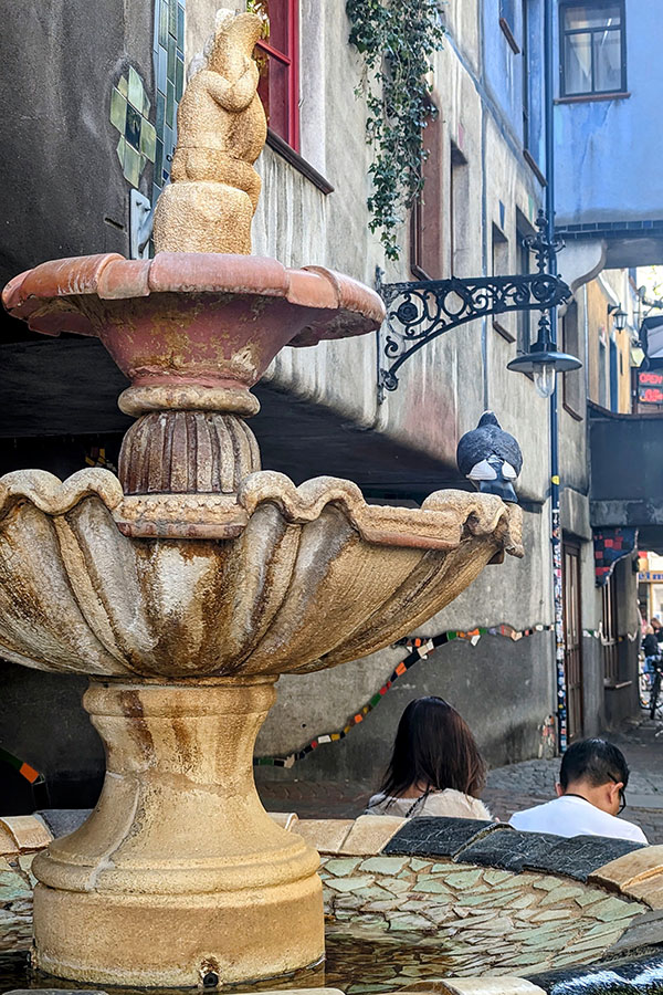 A whimsical fountain in Hundertwasser Village, Vienna, featuring a layered stone basin with a sculptural centerpiece. A pigeon perches on the edge, while colorful tiled details and the eclectic architecture of the village form the backdrop.