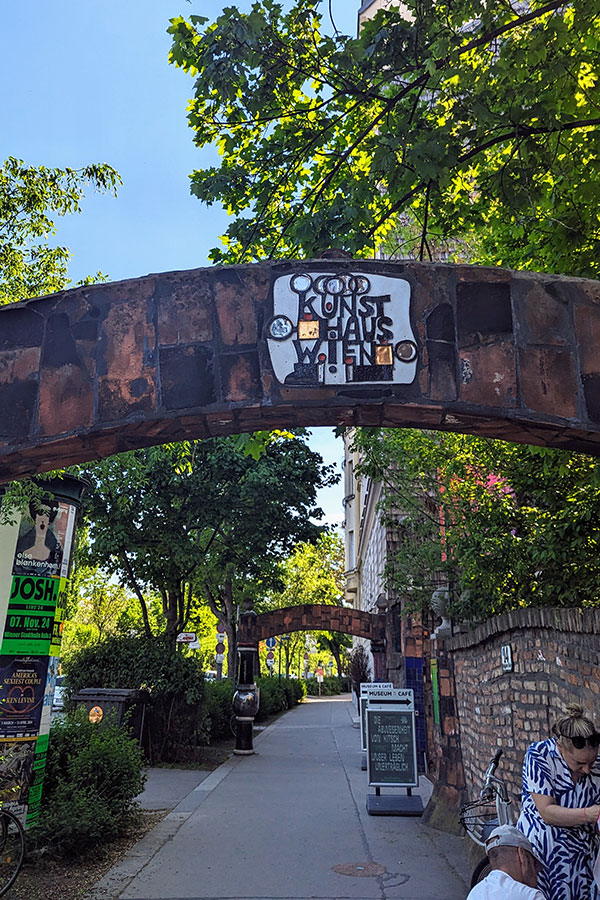 The artistic entrance gate to KunstHausWien in Vienna, featuring Hundertwasser's signature quirky style with asymmetrical shapes and earthy tones. The leafy trees and sunny pathway leading to the museum create a welcoming atmosphere.